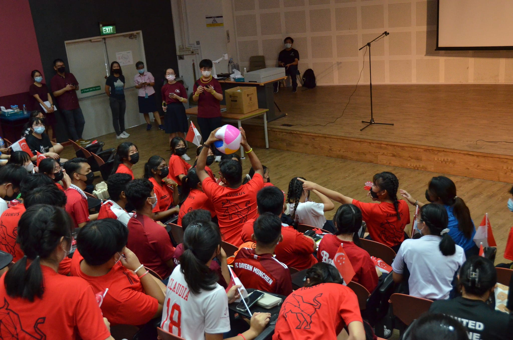 Students in red shirts sit in a hall as one holds up a colorful beach ball near a stage with a microphone stand.